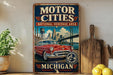 A vintage-style metal sign advertising the Motor Cities National Heritage Area in Michigan, featuring a classic red car and a bridge in the background.