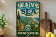 A vintage-style metal sign with the text "MOUNTAINS TO SEA TRAIL NORTH CAROLINA" displayed on a wooden shelf, with a scenic mountain landscape and a path leading to the sea in the background.