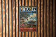 A vintage poster for Natchez National Historical Park, featuring a river scene with a steamboat and a house, is displayed on a wooden wall.