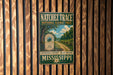 A vintage metal sign for the Natchez Trace National Scenic Trail, featuring a scenic image of a road and trees, mounted on a wooden wall.