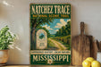 A vintage-style metal sign for the Natchez Trace National Scenic Trail, featuring a scenic landscape with a dirt road, trees, and a signpost.