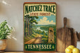 A vintage-style sign for the Natchez Trace State Forest in Tennessee, featuring a scenic landscape with a dirt road, trees, and a river.