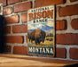 A vintage metal sign displaying the text "NATIONAL BISON RANGE MONTANA" with an illustration of a bison in a grassy field and mountains in the background.