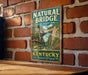 A vintage-style metal sign for Natural Bridge State Resort Park in Kentucky, featuring a scenic illustration of the bridge surrounded by lush greenery.