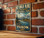 A vintage-style metal sign with a scenic mountain landscape and a river, mounted on a brick wall.