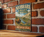A vintage-style metal sign for Niobrara Valley Preserve in Nebraska, featuring a buffalo in a grassy field with a river and mountains in the background.
