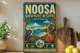 a vintage-style sign for the Noosa Bioscape Reserve in Australia, with illustrations of a koala, a bird, and a tropical landscape. The sign is displayed on a shelf next to a wooden cutting board and a potted plant.