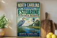 A vintage-style sign for the North Carolina Estuarine Research Reserve, featuring a blue heron and a lighthouse, is displayed on a shelf next to a wooden cutting board and some lemons.