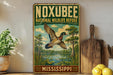 A vintage-style sign for the Noxbee National Wildlife Refuge in Mississippi, featuring a colorful illustration of a bird in flight over a pond surrounded by trees and greenery.