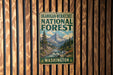A vintage-style metal sign for the Okanogan-Wenatchee National Forest in Washington, featuring a mountain landscape with a river and pine trees.