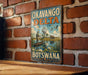 A vintage-style metal sign with the text "OKAVANGO DELTA SYSTEM" and "BOTSWANA" is displayed on a brick wall.