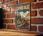 A vintage-style metal sign with the text "Old and New Towns of Edinburgh United Kingdom" and an illustration of a city street with a double-decker bus.