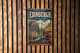 A vintage metal sign with the text "Old and New Towns of Edinburgh United Kingdom" and a picture of a city street with a red double-decker bus, mounted on a wooden wall.
