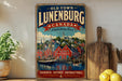 a vintage-style metal sign with a colorful illustration of a town, including buildings, boats, and a river, along with the text "OLD TOWN LUNENBURG CANADA A World Heritage Trust" and "COLOURFUL, HISTORIC, UNFORGETTABLE." The sign is displayed on a shelf next to a wooden cutting board and some potted plants.