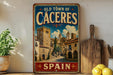A vintage-style metal sign with the text "Old Town of Cáceres" and "Spain" on it, featuring an illustration of a historic town with buildings and palm trees. The sign is displayed on a shelf next to a wooden cutting board and a potted plant.