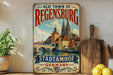 a vintage-style metal sign for the Old Town of Regensburg, Germany, with a painting of the town's landmarks and a river. The sign is displayed on a shelf next to a wooden cutting board and some potted plants.