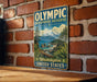 A vintage-style metal sign with the text "OLYMPIC BIOSPHERE RESERVE Washington United States" is displayed on a brick wall.