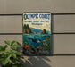a metal sign with a scenic ocean view, including a whale, a seagull, and a rocky coastline. The sign is mounted on a wall and is surrounded by greenery, including a plant.