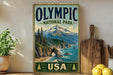 A vintage-style sign for Olympic National Park in the United States, featuring a scenic mountain landscape with pine trees and a beach.