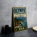 A vintage-style sign for Olympic National Park in the USA, featuring a scenic mountain landscape with pine trees and a body of water.