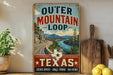 A vintage-style sign advertising the Outer Mountain Loop in Texas, featuring a scenic mountain landscape and a road winding through it.
