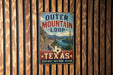 A vintage-style sign for the Outer Mountain Loop in Texas, featuring a scenic road surrounded by mountains and a river.