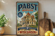 A vintage-style metal sign for Pabst Palace, a historic mansion in Milwaukee, Wisconsin, with a large illustration of the building and the text "Milwaukee's Brewing Heritage Wisconsin".