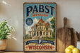 A vintage-style metal sign for Pabst Palace, a historic mansion in Milwaukee, Wisconsin, with a large illustration of the building and the text "Milwaukee's Brewing Heritage Wisconsin".
