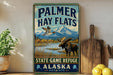 A vintage-style sign for Palmer Hay Flats, a state game refuge in Alaska, featuring a moose and a bird in a scenic landscape.
