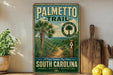 A vintage-style metal sign for the Palmetto Trail in South Carolina, featuring a palm tree and a trail marker, is displayed on a shelf next to a wooden cutting board and some lemons.