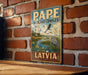 A vintage-style metal sign with the text "PAPE WETLAND COMPLEX LATVIA" is displayed on a brick wall.