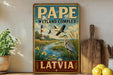 A vintage-style metal sign with a painting of a lighthouse, birds, and a pond, along with the text "PAPE WETLAND COMPLEX LATVIA".