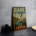 A vintage-style metal sign with the text "PAPE WETLAND COMPLEX" and "LATVIA" on it, featuring an illustration of a lighthouse and birds in a wetland landscape.