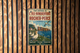 A vintage metal sign with text and an illustration of a coastal scene, mounted on a wooden wall.