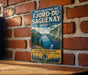 A vintage-style metal sign with a scenic mountain landscape and a bird in flight, mounted on a brick wall.