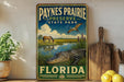 A vintage-style sign for "Paynes Prairie Preserve State Park" in Florida, featuring an illustration of a horse, alligator, and bird in a grassy field with a body of water.