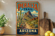 A vintage-style metal sign for the Peralta Trail in Arizona, featuring a desert landscape with mountains, cacti, and a winding path.