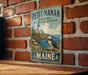 A vintage metal sign for the Maine National Wildlife Refuge, featuring a puffin bird and a lighthouse, is displayed on a brick wall.