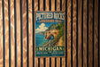 A vintage metal sign for the Pictured Rocks Lakeshore Trail in Michigan, featuring a scenic illustration of a rocky coastline with a path leading through the landscape.