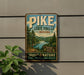 A vintage-style metal sign for Pike State Forest in Indiana, featuring a scenic landscape with pine trees, a river, and mountains.