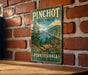 A vintage-style metal sign for Pinchot State Forest in Pennsylvania, featuring a scenic landscape with pine trees, mountains, and a lake.