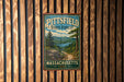 A vintage metal sign for the Pitsfield State Forest in Massachusetts, featuring a scenic landscape with pine trees, a lake, and mountains.