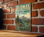A vintage-style metal sign for the Plumas National Forest in California, featuring a scenic mountain landscape with pine trees and a river.