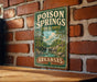 A vintage-style metal sign for the state of Arkansas, featuring a scenic illustration of a forest and a waterfall.