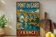 A vintage-style metal sign for the Pont du Gard in France, featuring a picturesque illustration of the aqueduct and the river below.