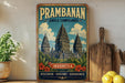 a vintage-style poster of the Prambanan Temple in Indonesia, with a wooden frame and a vintage-style design. The poster is displayed on a shelf next to a wooden cutting board and a potted plant.