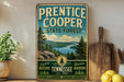 A vintage-style metal sign for Prentice Cooper State Forest, featuring a scenic mountain landscape and pine trees, is displayed on a shelf next to a wooden cutting board and some lemons.