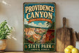 A vintage-style sign for Providence Canyon, Georgia, featuring a scenic landscape with mountains and a blue sky.
