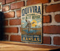 A vintage-style sign for the Quivira National Wildlife Refuge in Kansas, featuring a duck flying over a grassy field with a windmill in the background.