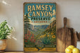 a vintage-style poster of Ramsey Canyon Preserve, Arizona, with a scenic mountain landscape and desert vegetation. The poster is displayed on a wall, and there are some kitchen utensils, such as a cutting board and a knife, as well as some potted plants and lemons in the foreground.